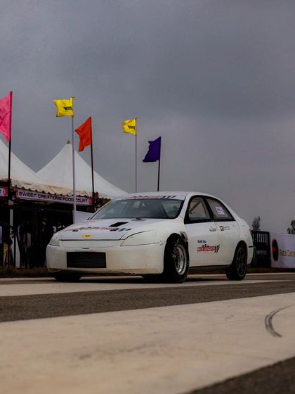 A white modified sedan at the starting line, ready to launch. The flags and tents in the background paint a picture of the full festival environment.