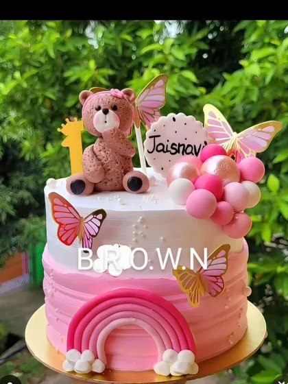 A sweet first birthday cake combining a teddy bear, butterflies, and a rainbow in a lovely pink ombre design.