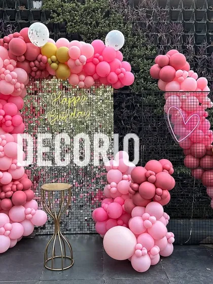 An artistic outdoor balloon installation in various shades of pink and gold against a silver sequin backdrop. The asymmetrical design creates a modern and high-fashion look.