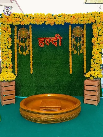 A simple yet beautiful Haldi decoration with a green grass backdrop, a frame of marigold flowers, and a neon Hindi "Haldi" sign. The wooden crates and dreamcatchers add a rustic, bohemian touch to the setup.