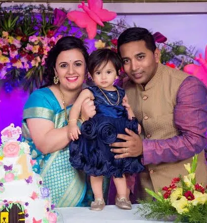 A family of three poses behind their daughter's first birthday cake. The parents are dressed in elegant traditional Indian wear, coordinating with their daughter's beautiful blue dress.