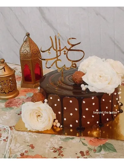 An elegant chocolate drip cake for Eid Mubarak. It's decorated with white polka dots, fresh white roses, and a beautiful Arabic calligraphy cake topper.