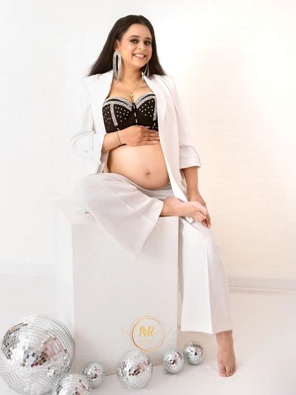 A joyful and relaxed portrait. The expectant mother smiles warmly, seated in a chic white suit on a studio block, surrounded by fun disco ball props.