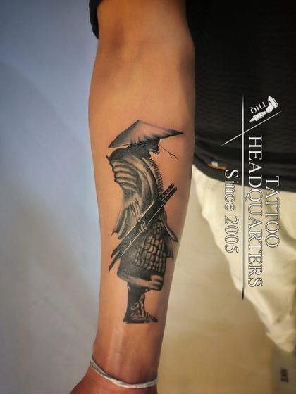 A stoic samurai warrior tattoo on the forearm. The black and grey shading creates a sense of depth and texture on the armor and helmet.