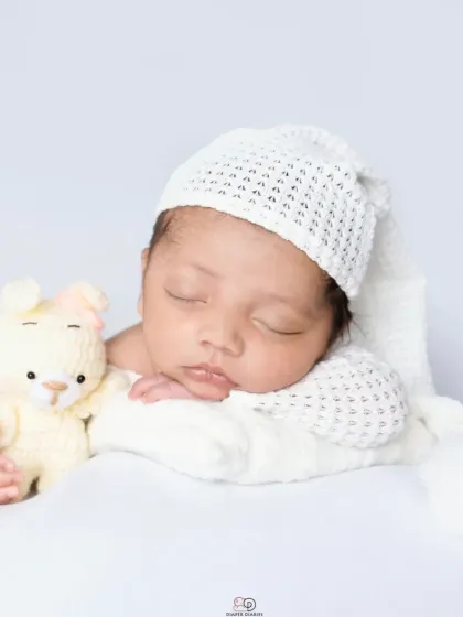 A pure and simple newborn portrait against a clean white background. The baby is dressed in a white knitted outfit and sleepy hat, showcasing their angelic features.