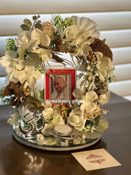 This darbar features a classic white and green floral arrangement around Guruji's photo, set on a mirrored base that reflects the light beautifully. A silver Om symbol and a tea light complete the sacred setting.