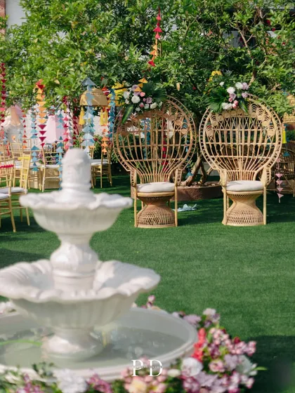 A Pinterest-perfect Mehendi setup in a garden, featuring peacock chairs for the couple, a beautiful white fountain, and colorful hanging decorations.