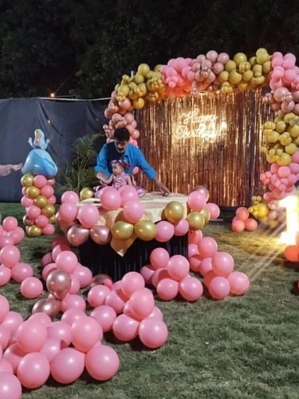 A beautiful outdoor first birthday party in a garden. The family is seen around a cake table framed by a pink and gold balloon arch against a sequin backdrop.