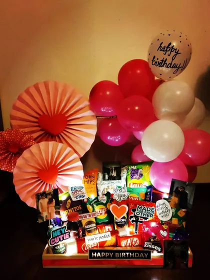This birthday hamper uses a vibrant pink and red color scheme with paper fan decorations and balloons. It's filled with snacks and photos, all illuminated by fairy lights for a festive evening celebration.