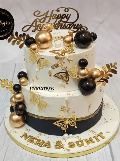 A luxurious two-tier choco-vanilla anniversary cake. The design features a black and gold theme with golden butterflies, decorative balls, and a custom nameplate.