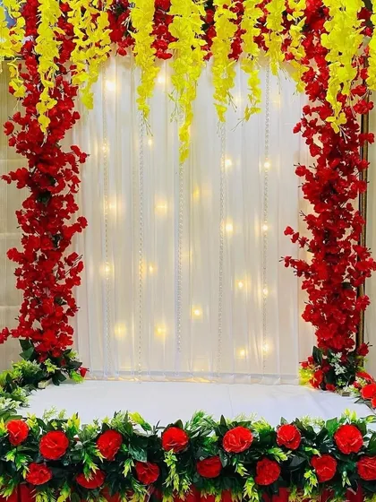 A vibrant red and yellow floral arch against a white curtain with fairy lights. The base is lined with red roses and greenery, creating a bold and festive look.