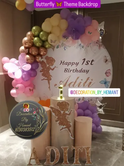 A beautiful butterfly-themed backdrop for a first birthday. The setup includes a personalized circular backdrop, pastel balloon garlands, and glittery fairy cutouts. The lit-up '1' and name letters add a special touch.