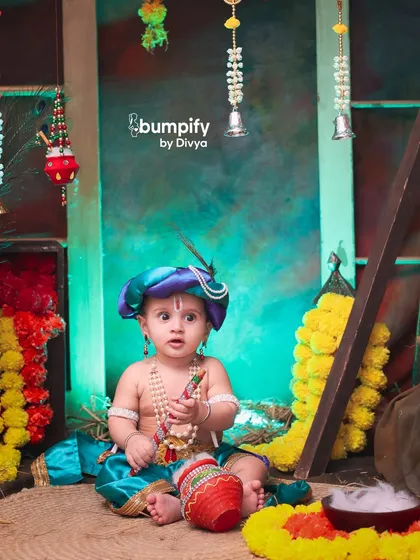 This little Krishna is sitting amidst a rustic, village-themed setup, complete with marigold garlands and a pot of butter, looking absolutely adorable.