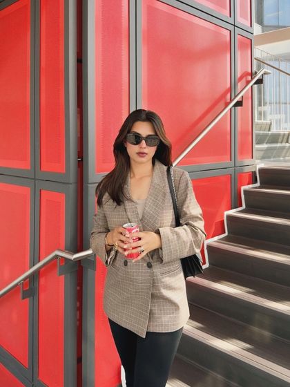A confident pose against a bold, red architectural background. This shot highlights a chic fall outfit, featuring a plaid blazer and sunglasses, perfect for a fashion brand's lookbook.