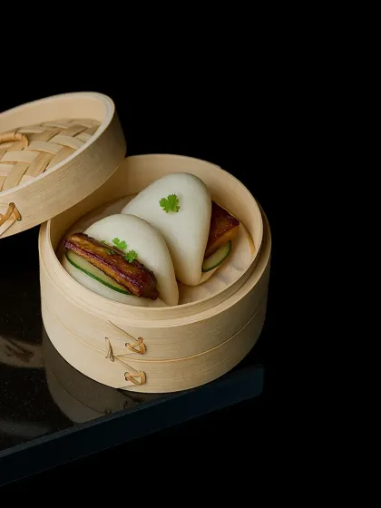 Our BBQ Pork Bao features delectable steamed buns filled with roasted pork, crisp cucumber slices, and our house BBQ sauce, served in a traditional bamboo steamer.