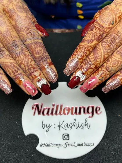 A rich and warm bridal nail set. This design combines deep red and pink nails with glitter accents and rhinestone bows, perfectly matching the intricate mehendi.