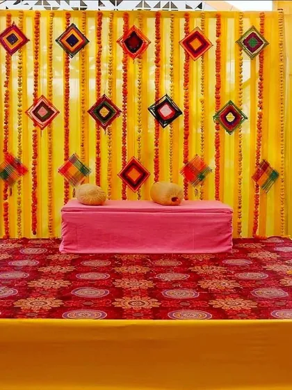 A classic Haldi or Mehndi stage with a backdrop of yellow drapes and marigold strings. The simple seating on a patterned rug is perfect for a traditional home ceremony.