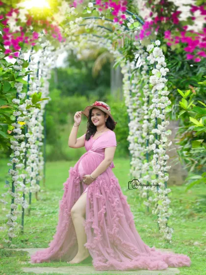A beautiful solo portrait under a floral archway. The mother-to-be is wearing a pink ruffled gown and a hat.