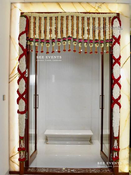 A pristine white marble pooja room entrance adorned with traditional red and white floral garlands. The contrast creates a very elegant and divine look.