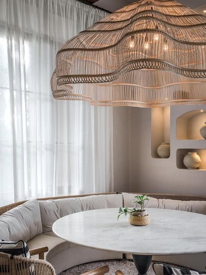 This intimate dining nook at EVE Bombay features a plush, curved banquette and a large, sculptural rattan chandelier. The design uses soft, ambient lighting and a neutral palette to create a warm and inviting atmosphere for guests.