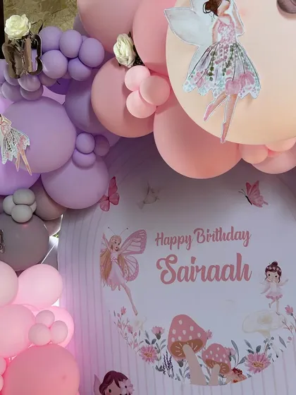 A close-up on the personalized "Happy Birthday Sairaah" backdrop, framed by a cascade of pink and purple balloons. This view highlights the adorable fairy and mushroom illustrations that define the enchanted forest theme.