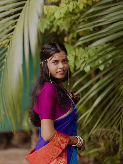 A beautiful portrait of a Maharashtrian bride in a vibrant blue and purple saree. Her simple yet elegant headpiece and joyful expression are perfectly framed by lush green leaves.