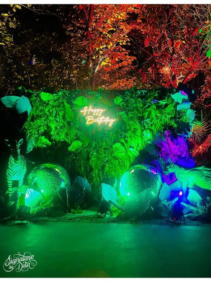 A wider shot of the neon photo wall, showing how the installation is set against the natural backdrop of the trees, blending the artificial and natural elements.