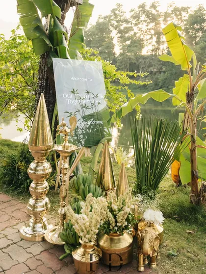 The entrance to the lakeside South Indian wedding, with a clear acrylic welcome sign set amongst brass decor, banana trees, and lush florals, perfectly blending modern and traditional elements.