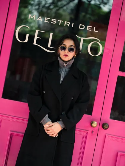 A full-body shot of a model in a black trench coat and sunglasses, posing against a bright pink door. The color contrast and confident stance create a bold street-style statement.