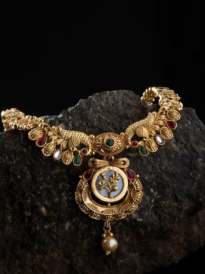A traditional temple jewelry necklace featuring intricate carvings. The piece is laid on a dark, textured rock to emphasize its antique gold finish and detailed workmanship.