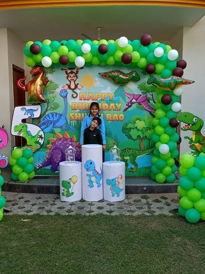 A lively dinosaur-themed birthday party in a backyard. This setup includes a large backdrop with various dinosaurs, a green balloon arch, and themed pedestals for cake and props.