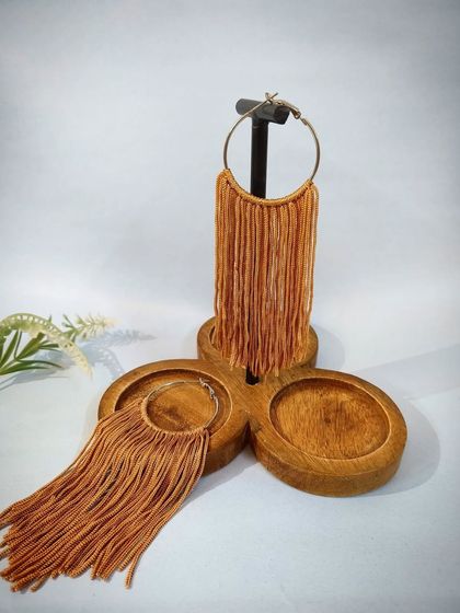 A product shot of the peach-brown tasselled hoop earrings, displayed on a wooden stand.