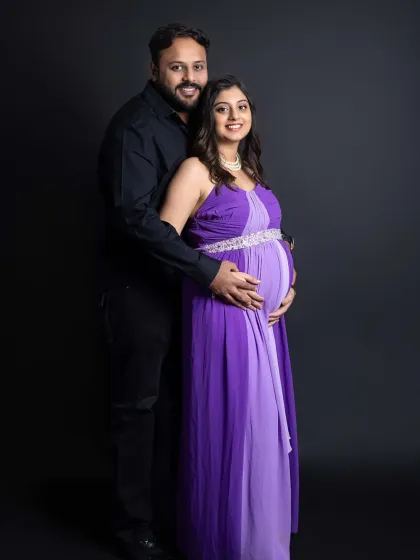 A classic maternity couple portrait. The parents-to-be stand together, smiling at the camera, showcasing their coordinated formal outfits against a timeless dark studio background.
