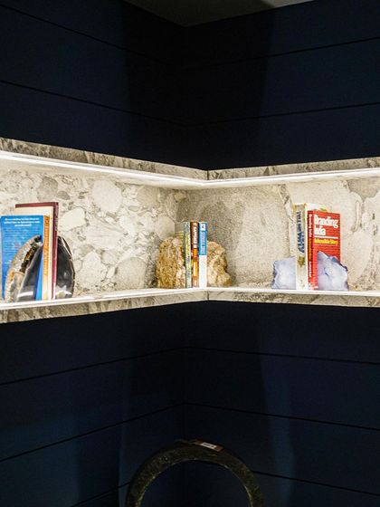 This custom-designed, L-shaped marble bookshelf is seamlessly integrated into a corner, with built-in lighting that casts a soft glow. Set against the deep blue grooved wall, it stands out as an elegant and functional centerpiece.