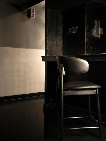 A solitary bar stool, hinting at the quiet, personal moments the space is designed to foster. The signage for "Sushi & Sake" is subtly integrated into the raw concrete wall.