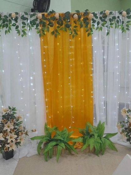 A simple yet elegant backdrop for a home function. This design uses white and yellow drapes with fairy lights, complemented by artificial plants and flowers for a fresh look.