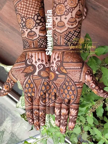 A close-up of a heavy Indian design showcasing twin peacocks on each palm. The fingers are adorned with lotus motifs, and the arms feature bands of checks and hanging jhumkas.