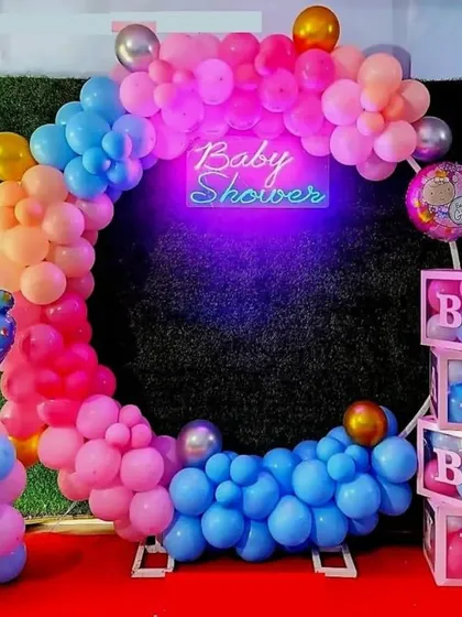 A vibrant and colorful baby shower backdrop. The rainbow-colored balloon ring against a green turf wall creates a stunning photo booth, complete with a neon sign and 'BABY' blocks.