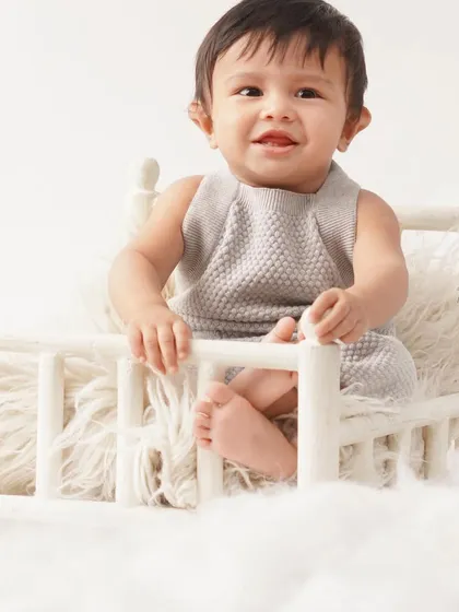 A charming baby boy in a grey knit romper sits on a miniature white bed, surrounded by soft, cloud-like fluff for a dreamy portrait.