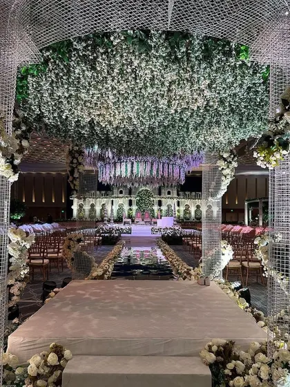 We create breathtaking entrances that transport guests into another world. This walkway, framed by a modern lattice structure and a ceiling of lush white and green florals, sets the stage for an enchanted garden wedding. 💚