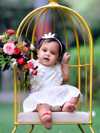 A sweet baby girl in a white dress, sitting in a golden birdcage prop and holding a bouquet of flowers during an outdoor session.