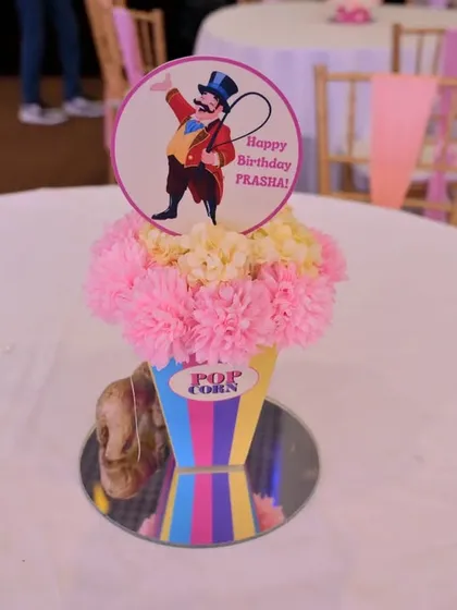 A custom circus-themed centerpiece, designed to look like a box of popcorn filled with pink flowers, topped with a personalized sign for the birthday girl.