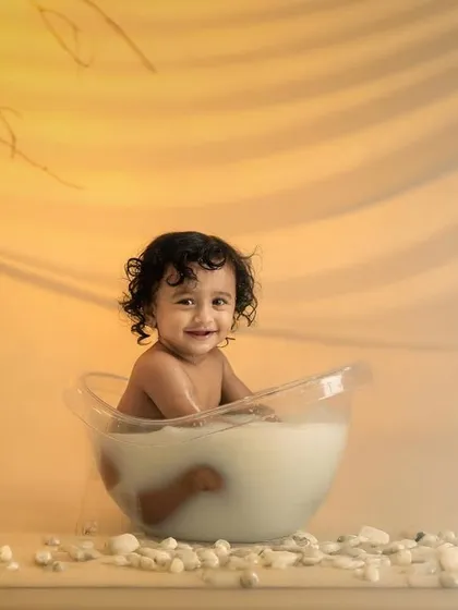 A smiling toddler enjoys a fun milk bath in a clear tub, set against a warm, glowing yellow background.