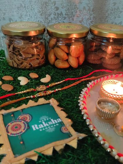 A premium Rakhi hamper setup showing three glass jars of my homemade Ragi Chips, Khasta Kachori, and Ragi Cookies, alongside a traditional pooja thali.