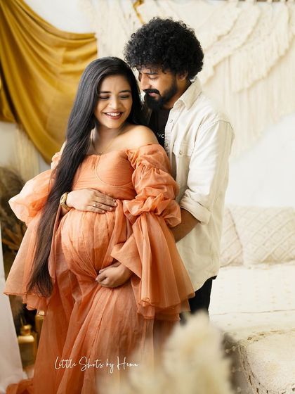 A candid and happy moment between a couple. The partner embraces the mom-to-be, who is dressed in a flowing peach gown, as they share a smile in a boho-styled room.
