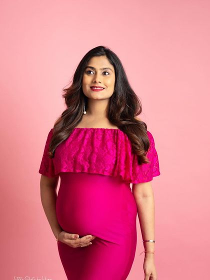 A close-up portrait of a mother-to-be in a vibrant pink dress. She looks up thoughtfully against a matching pink background, her hand gently resting on her bump.