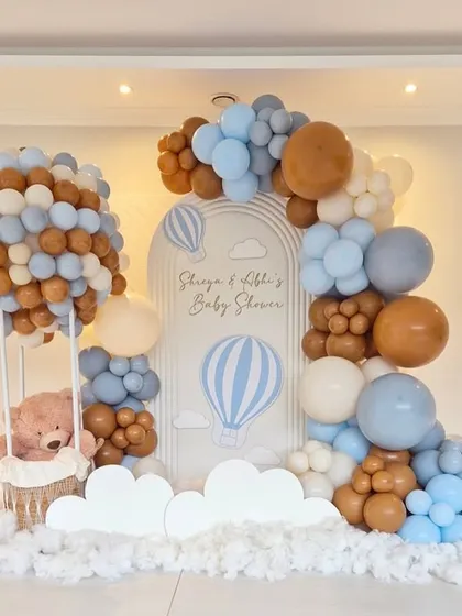 An elaborate 'Up, Up and Away' hot air balloon themed baby shower. This setup includes a large balloon sculpture forming a hot air balloon with a basket for a teddy bear, set against a custom backdrop and surrounded by clouds of balloons.