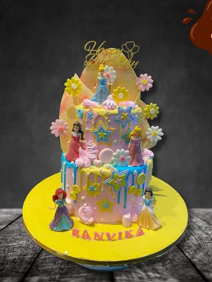 A dreamy two-tier cake featuring several Disney princesses. The pastel colors, star decorations, and drip effect create a magical backdrop for figures of Cinderella, Ariel, and others.