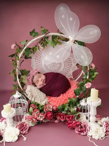 A magical, fairytale scene. This wide shot shows the newborn sleeping in a floral hoop with butterfly wings, surrounded by candles and birdcages for an enchanted forest feel.