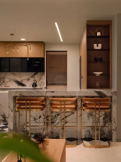 A view from the living area into the kitchen, showing the marble bar counter with stylish wooden and brass stools. An open shelving unit provides space for display.